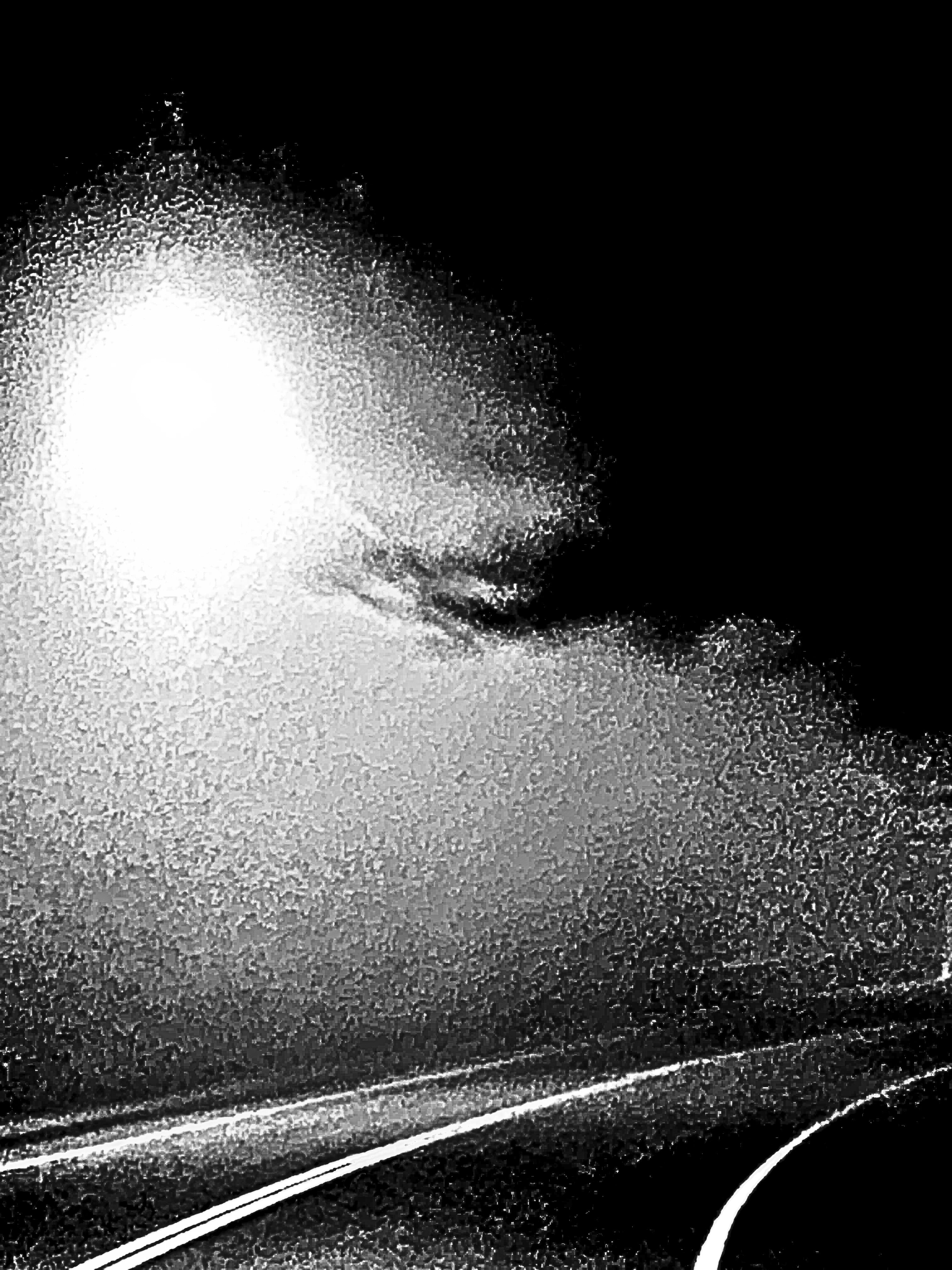 A black and white road curved towards the right lit under moonlight within a cloud of great fog.
